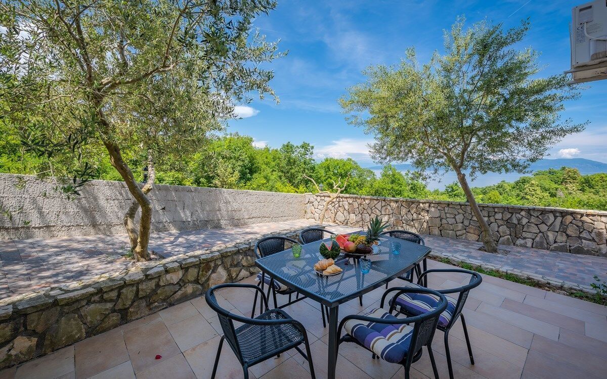 Eine Terrasse im Freien mit Tisch und Stühlen, umgeben von Steinmauern und Bäumen, mit Blick auf eine üppige Landschaft.