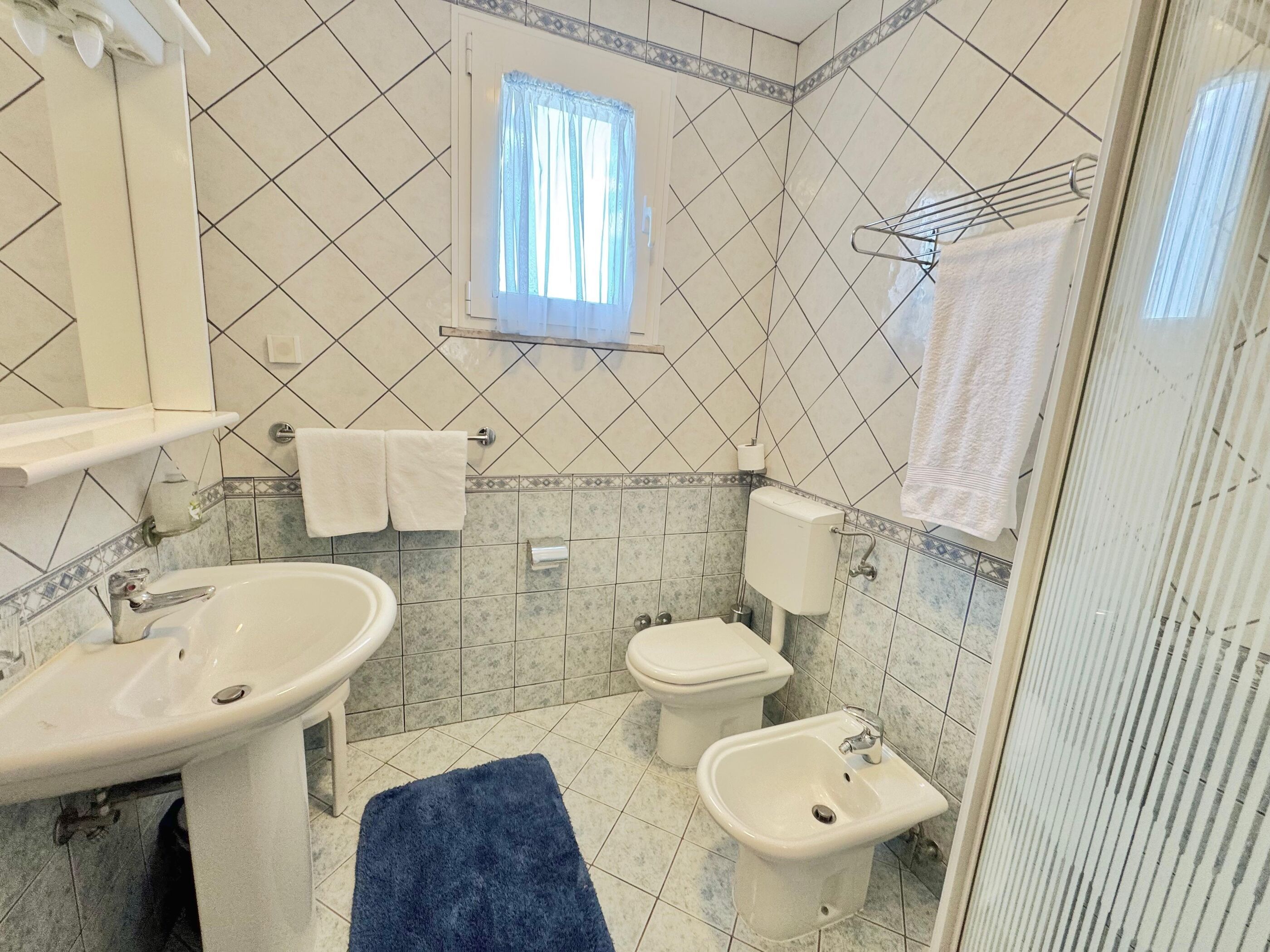 Ein Badezimmer mit Waschbecken, Bidet und Toilette, mit gefliesten Wänden und einem Fenster mit Vorhängen.