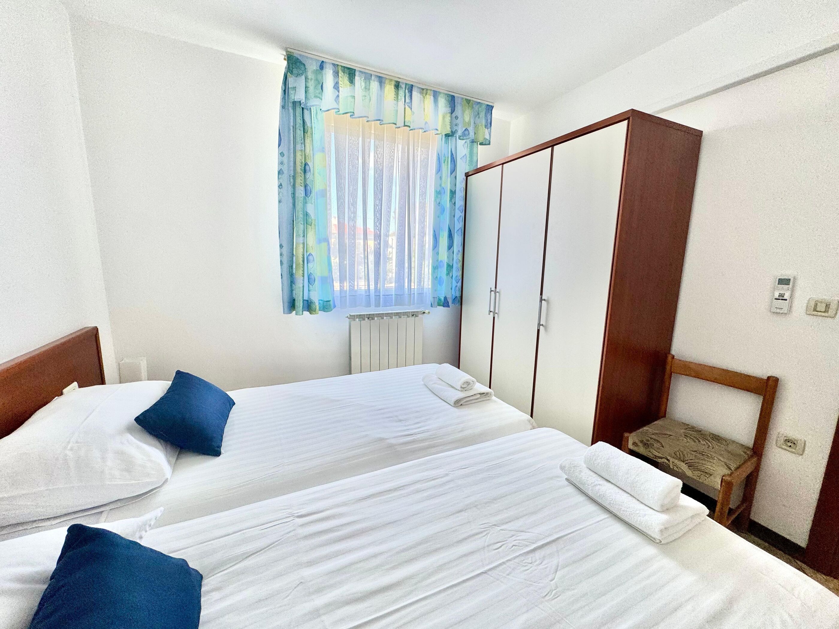 Habitación de hotel con dos camas, un armario de madera, una ventana con cortinas y un radiador.