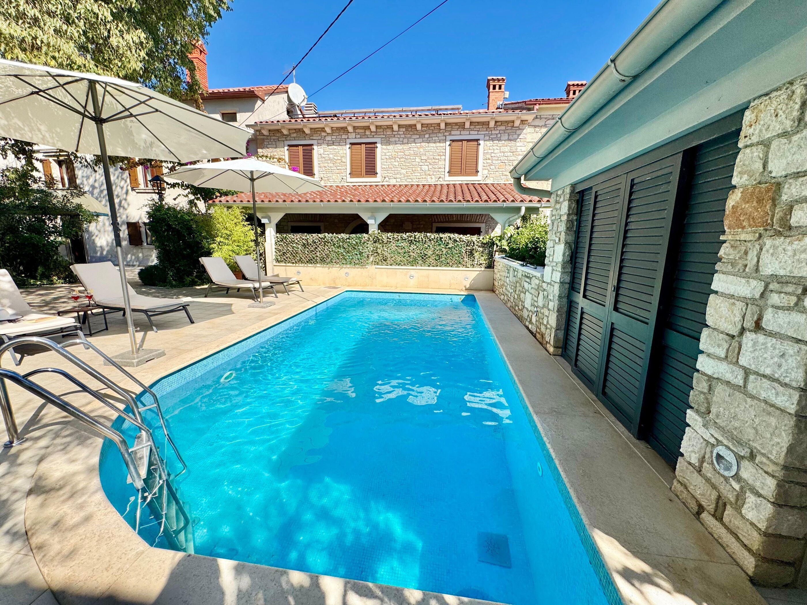 Une piscine entourée de chaises longues et de parasols, elle-même bordée de murs en pierre et d’un bâtiment doté d’un toit en tuiles.