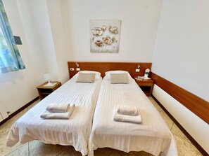 Une chambre d’hôtel avec deux lits, chacun recouvert d’une courtepointe blanche et agrémenté d’une serviette pliée. On y trouve une table de chevet avec une lampe, ainsi qu’une petite table sur laquelle sont posés un livre et un vase avec une fleur. Un tableau est accroché au mur, au-dessus des lits.
