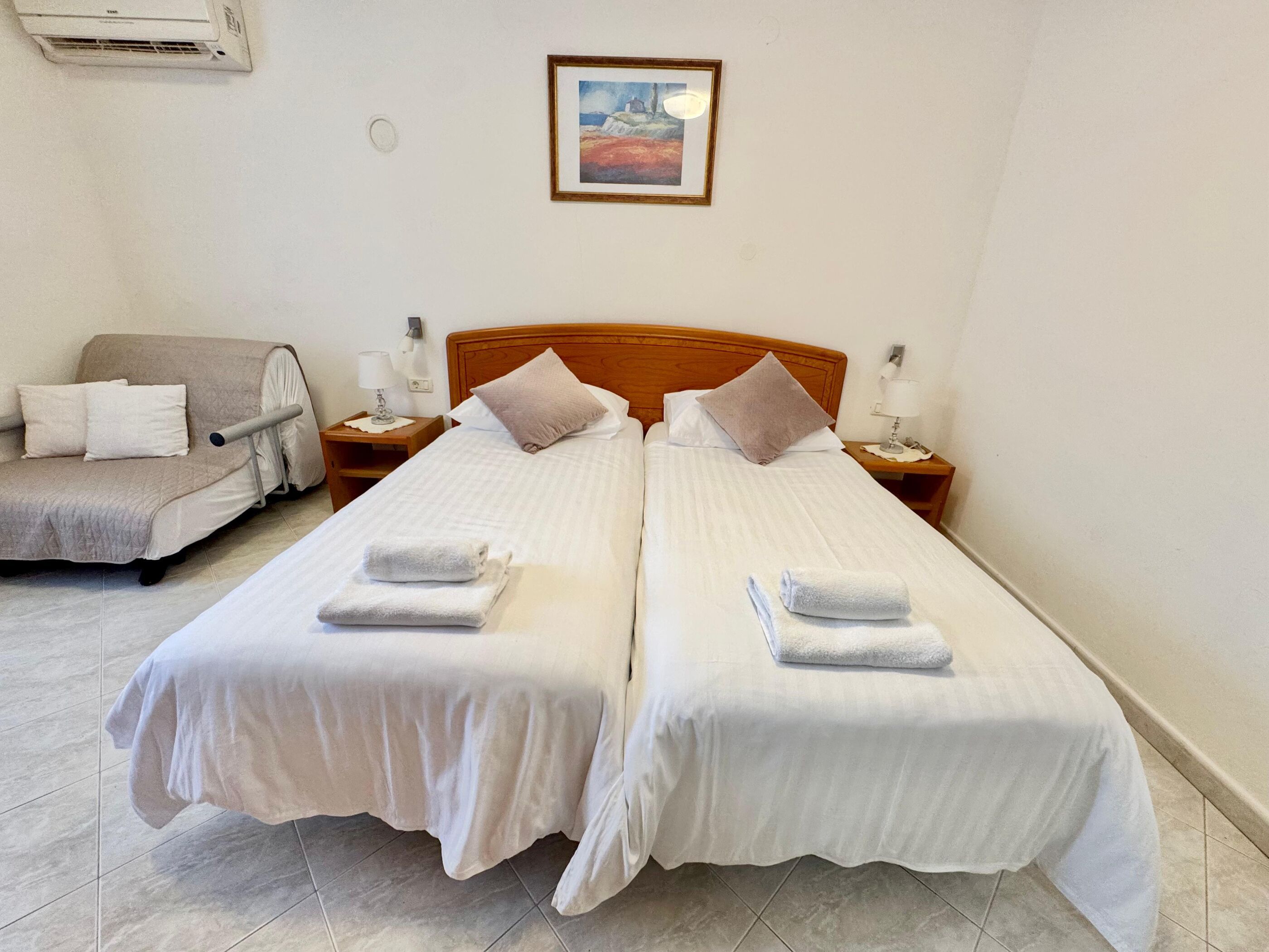 Habitación doble con dos camas, cada una con cubrecama blanco y dos almohadas. Tiene cabecera de madera, mesita de noche con lámpara y un cuadro en la pared.
