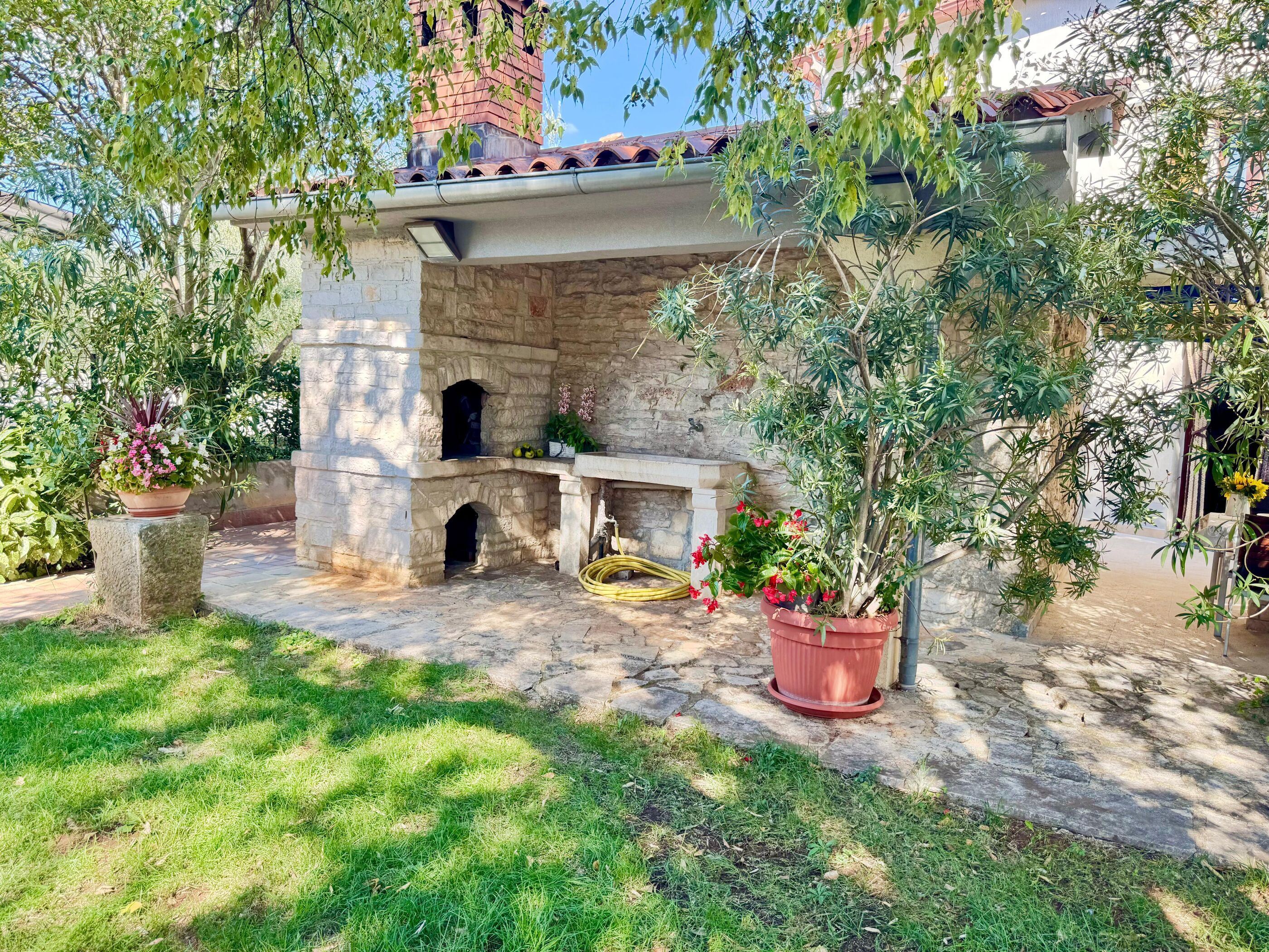 A stone-built outdoor kitchen area with a chimney, surrounded by potted plants and a grassy lawn.
