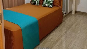 Ein Zimmer mit einem Einzelbett, einer braunen Bettdecke, zwei Kissen mit grünem Blattmuster, einer türkisblauen Zudecke und einem weißen Nachttisch mit Spiegel.
