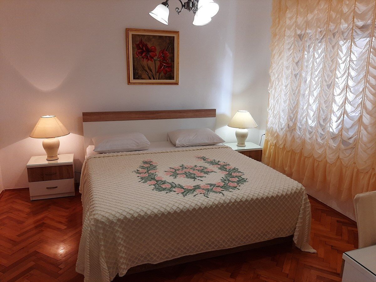 Ein Schlafzimmer mit Holzboden, einem Bett mit blumigem Überwurf, zwei Nachttischlampen, einem Nachttisch, einem gerahmten Bild an der Wand und einem durchsichtigen Vorhang am Fenster.