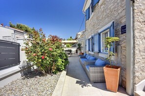 Une rue étroite bordée d’un immeuble en pierre, avec des volets bleus et une enseigne indiquant qu’il s’agit d’un appartement. On y trouve un petit jardin avec un arbuste et une plante en pot.