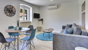Uma sala de estar moderna com um sofá cinzento, cadeiras azuis, uma mesa de madeira redonda e um relógio na parede.