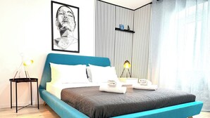 Quarto moderno com cama forrada em azul, mesa de cabeceira com abajur, obra de arte emoldurada na parede e painel de parede com listras verticais.