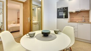 Una cocina compacta con electrodomésticos modernos, una zona de comedor con una mesa redonda y sillas, y un dormitorio visible con una cama y una mesita de noche.