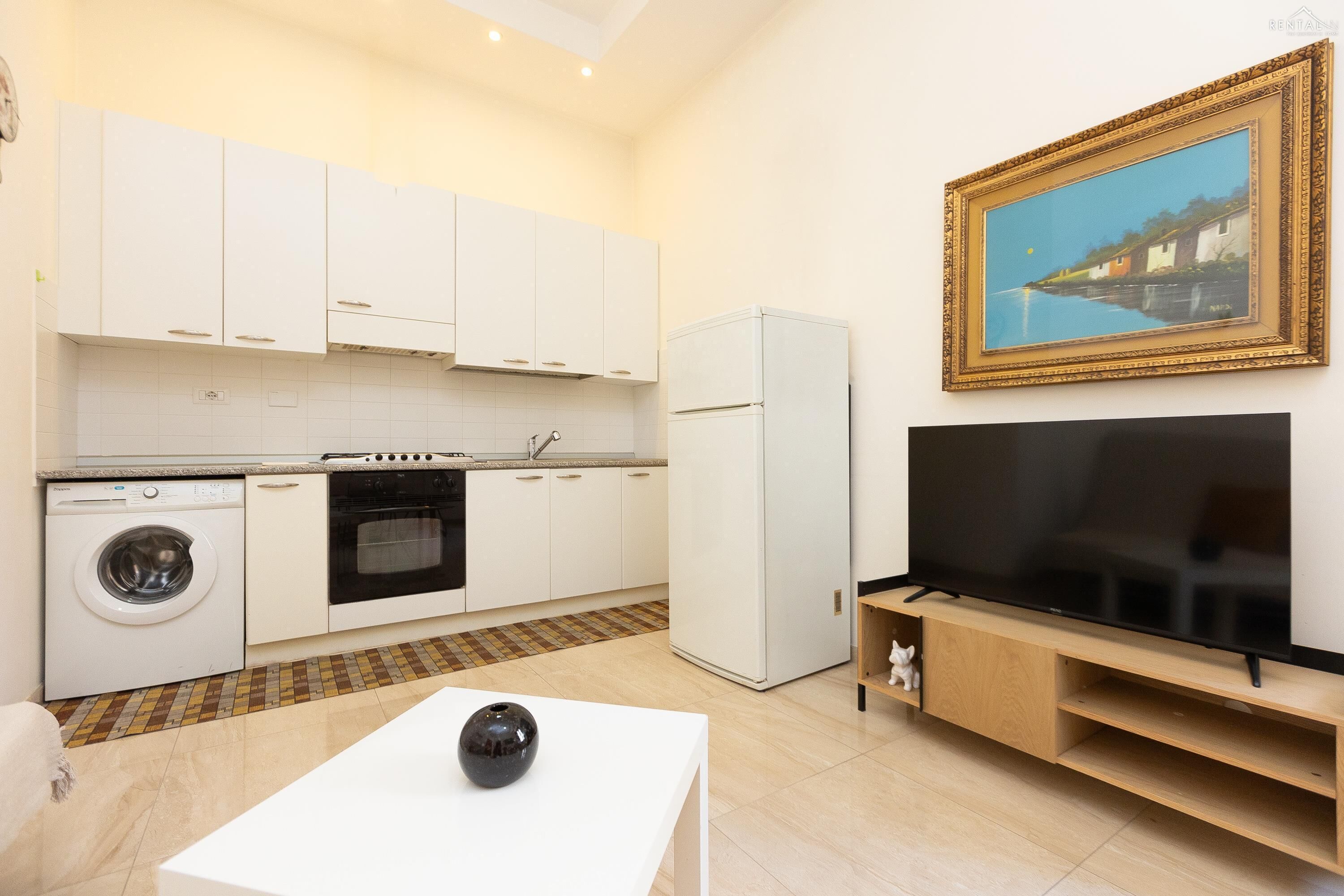 Una cocina moderna con armarios blancos, lavadora, horno y refrigerador. Hay un televisor de pantalla plana sobre un atril de madera y un cuadro en la pared.