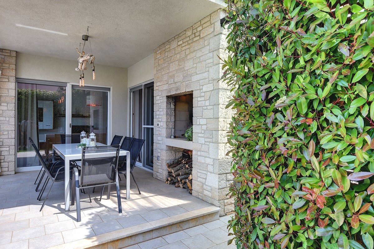 An outdoor patio with a stone fireplace, a dining table with chairs, and a hedge.