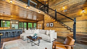 A spacious living area with a wooden interior, a sofa set, a coffee table, and a staircase with a metal railing. - Riverstone Estate | Luxe Creekside Group Getaway (Hochatown)