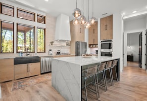 A modern kitchen with a large island, stainless steel appliances, and a view of trees through large windows. - Riverstone Estate | 4 Separate Cabins, Sleeps 66 (Hochatown)