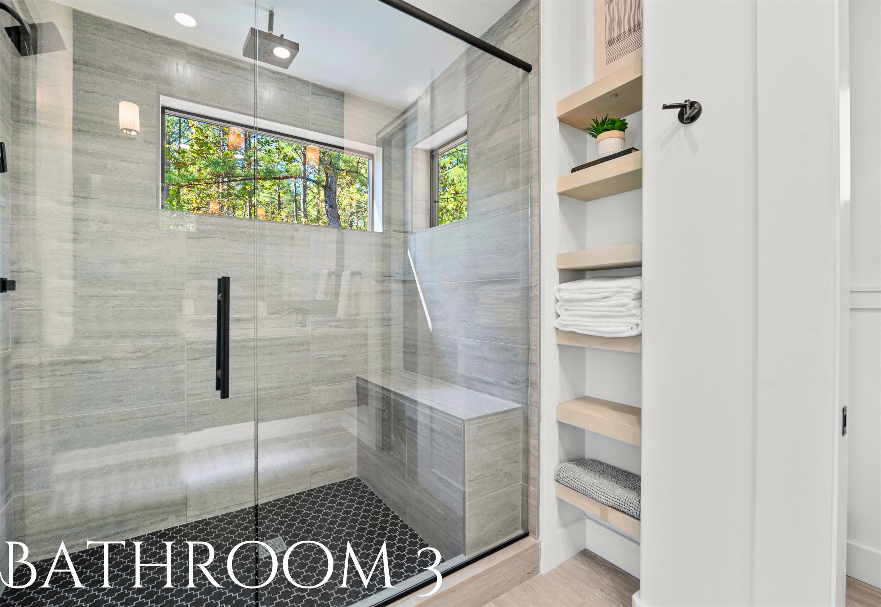 Un baño moderno con cabina de ducha de vidrio, banco incorporado y ventana con vistas a árboles.