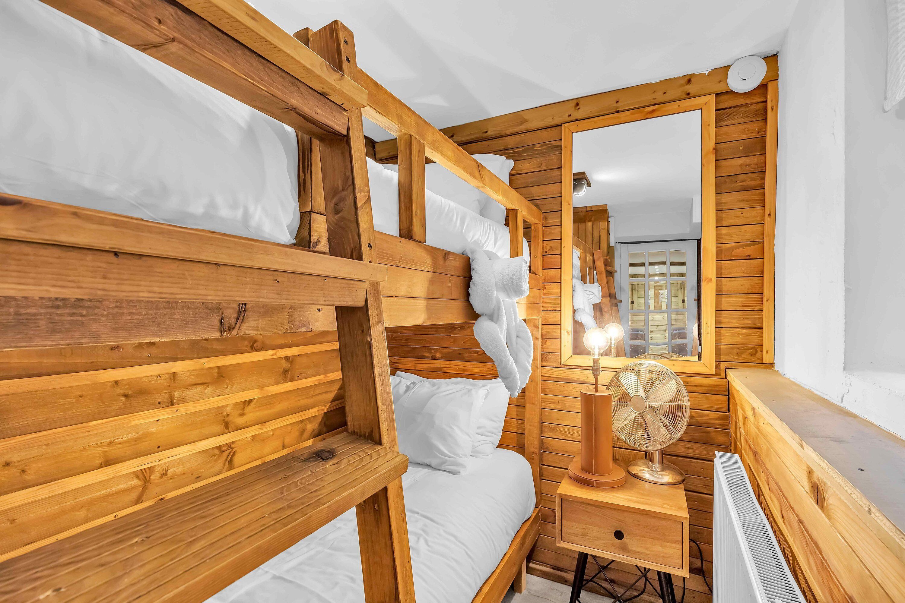 A wooden bunk bed with white bedding, a bedside table with a fan, and a mirror on the wall.