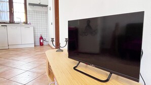 A flat-screen TV on a wooden stand in a kitchen with a tiled floor, a fire extinguisher, and a window with curtains. - Apartment with private parking just a 10-minute walk from the historic center (Lucca)