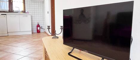 A flat-screen TV on a wooden stand in a kitchen with a tiled floor, a fire extinguisher, and a window with curtains.