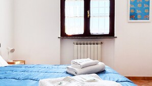 A neatly made bed with blue bedding and white towels, a radiator, and a window with a view of the outside. - Apartment with private parking just a 10-minute walk from the historic center (Lucca)