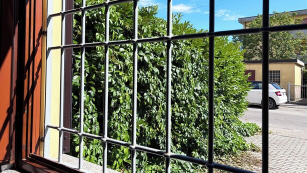 A window with metal bars, a view of a white car parked outside, and a building with a yellow facade in the background. - Apartment with private parking just a 10-minute walk from the historic center (Lucca)