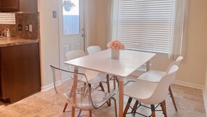 A dining area with a white table and chairs, a clear glass chair, a vase with flowers, a wooden cabinet, and a window with blinds. - Clean & Calm 3BR Home Near Houston Premium Outlets (Cypress)