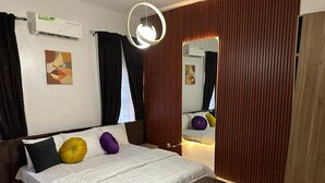 A hotel room with a bed, a bedside table, a mirror, a wall-mounted air conditioner, and a window with curtains. - 3 Bedroom En-Suite, Wi-Fi, Secured, 24/7 Power (Lekki)