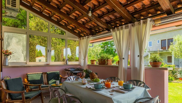 Une terrasse couverte avec une table dressée pour le petit-déjeuner, entourée de chaises, et donnant sur un jardin.