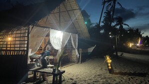 A beachside hut with a thatched roof, a table with a chair, and a small table with a plant and a teapot.