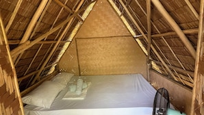 A small, single-bed room with a bamboo ceiling and walls, a bed with a white mattress and pillows, and a green fan.