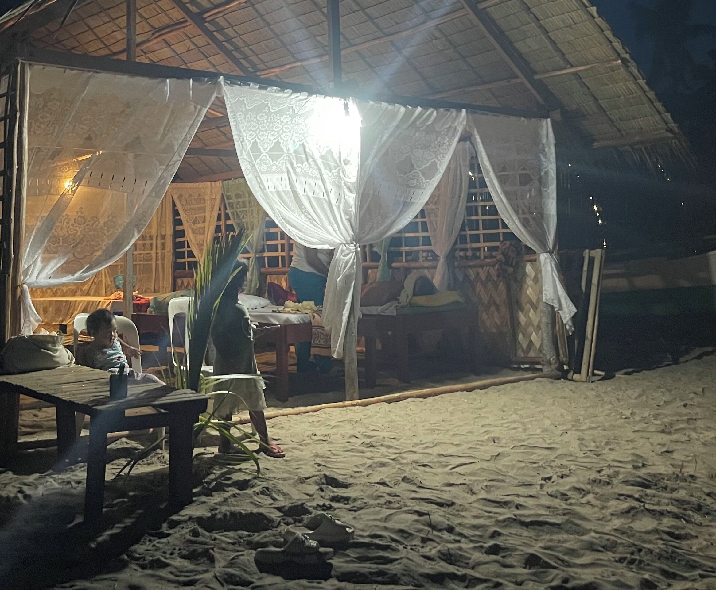 Una cabaña junto a la playa con techo de paja, una cama con mosquitero y una mesa con sillas.
