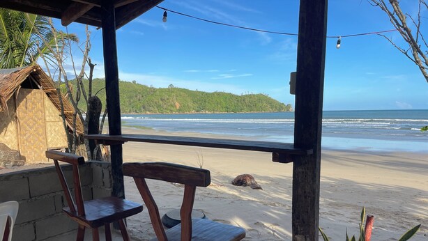 A beachside wooden structure with two chairs and a table, overlooking a sandy beach and ocean.