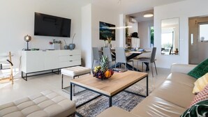 Un saló modern amb un sofà, una taula de centre amb fruita, una televisió i una zona de menjador amb caires i una taula.