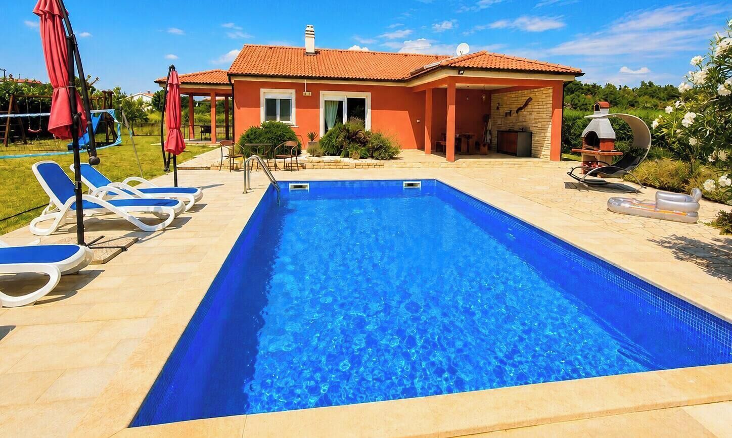 A swimming pool with lounge chairs, a barbecue area, and a house with a tiled roof.