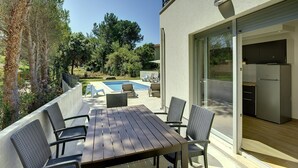 Patio esterno moderno con tavolo e sedie in legno, piscina e cucina visibile attraverso porte in vetro.