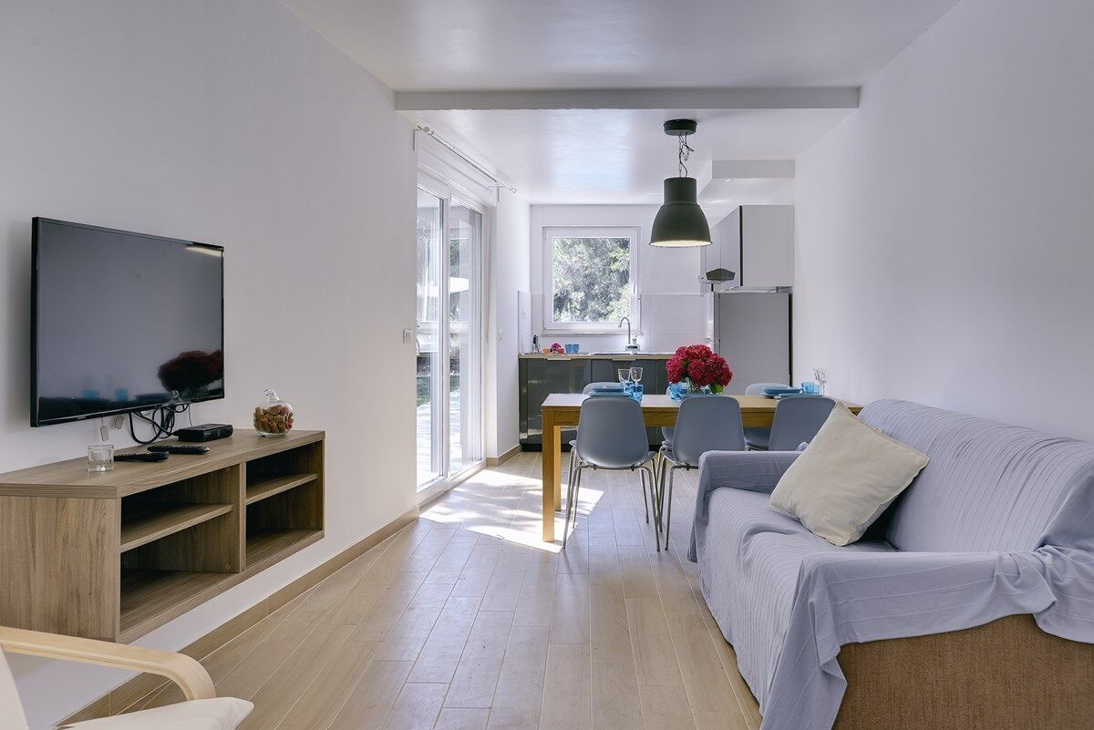 A modern living room with a flat-screen TV, wooden entertainment unit, grey sofa, dining table with chairs, and a kitchen area with a window.