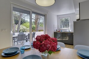 Un espace repas moderne avec une table dressée pour deux personnes, un vase de fleurs et une vue sur l’extérieur grâce à de grandes portes coulissantes.