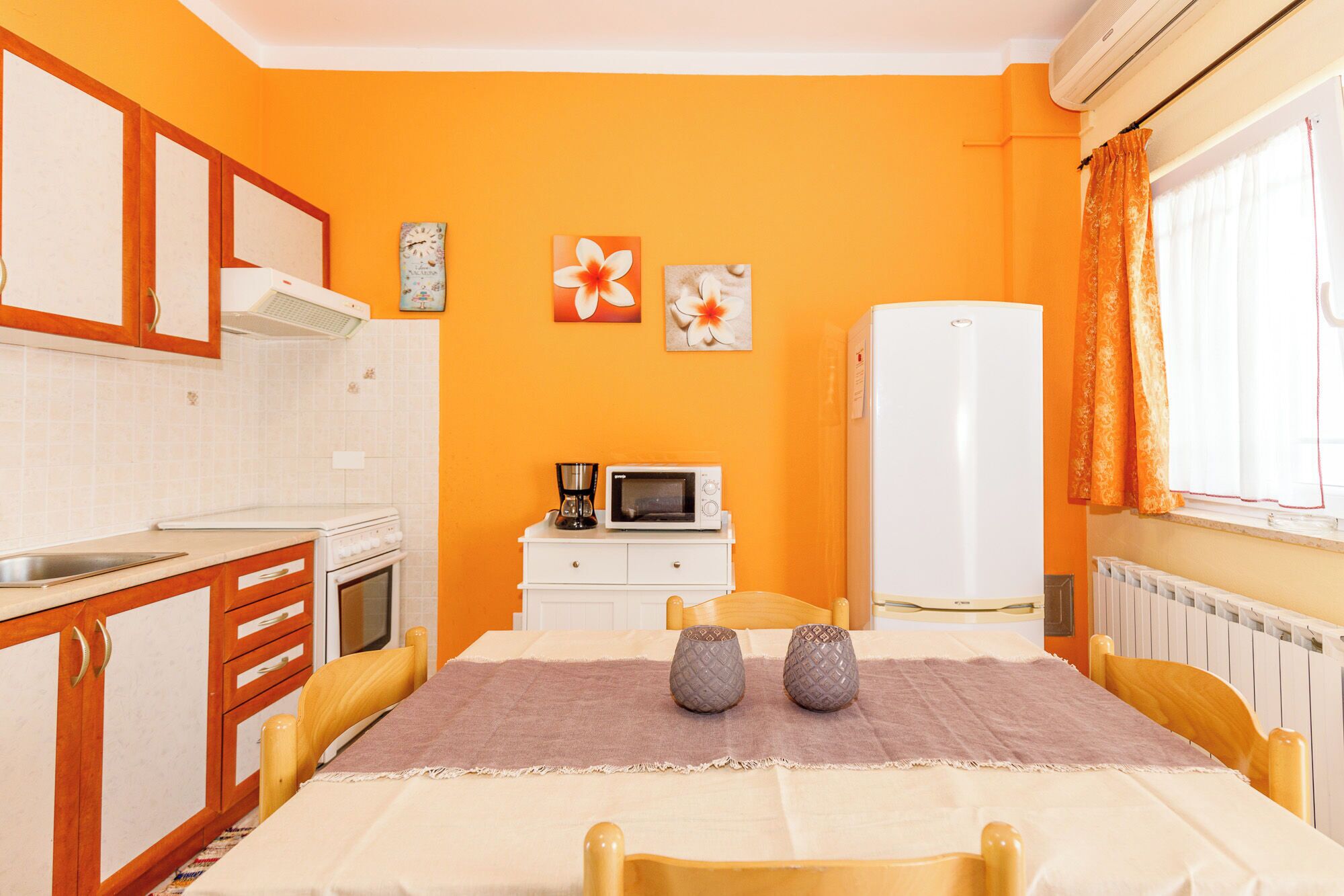 A compact kitchen with a microwave, stove, and refrigerator. There is a dining table with two chairs and a tablecloth.