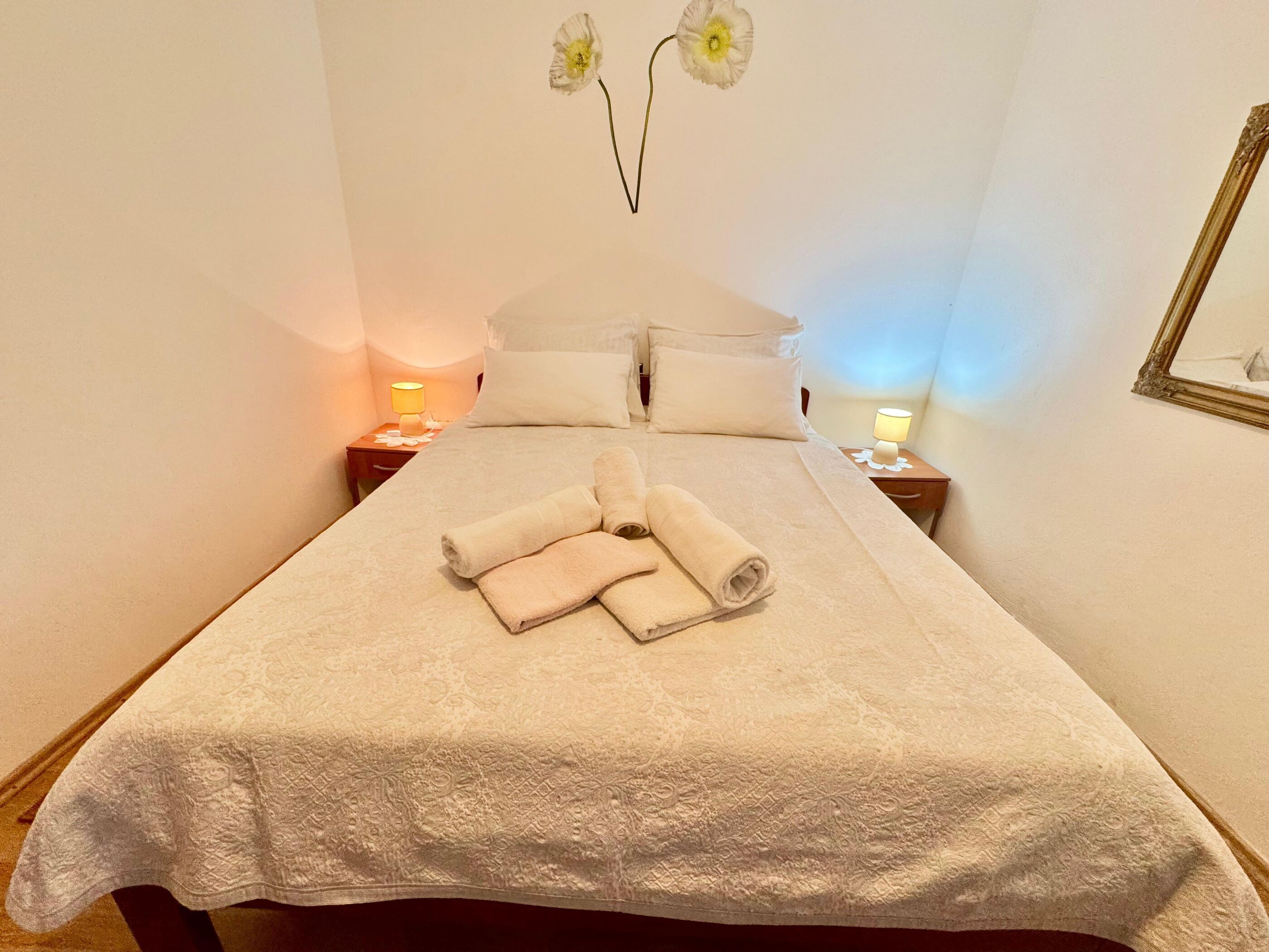 Habitació d'hotel amb un llit, dues taules de nit amb làmpades, un mirall fixat a la paret i un ram de flors.