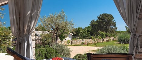 A covered outdoor seating area with wicker furniture, a table with a plate of strawberries, and a view of a stone wall and greenery.