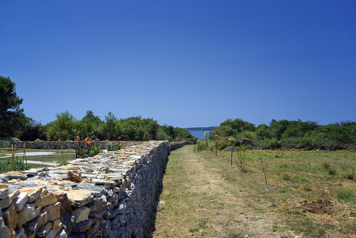 Un muro di pietra con un sentiero che conduce a una distesa d'acqua, circondato da vegetazione e un cielo sereno.