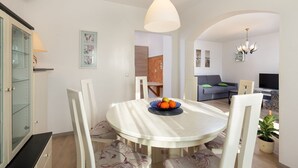 Een eetruimte met een ronde tafel, witte stoelen en een schaal fruit. Achtergrond heeft een bank en een televisie.