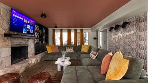 A cozy living room with a stone fireplace, a sectional sofa, and a flat-screen TV displaying a streaming service interface. - 5BR Oasis w Pool, Spa & Fire Pit (Round Rock)
