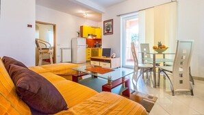 Uma sala de estar moderna com uma mesa de centro de vidro, um sofá com almofadas listradas, uma área de jantar com uma mesa e cadeiras de vidro, e uma cozinha com armários amarelos e uma geladeira branca.