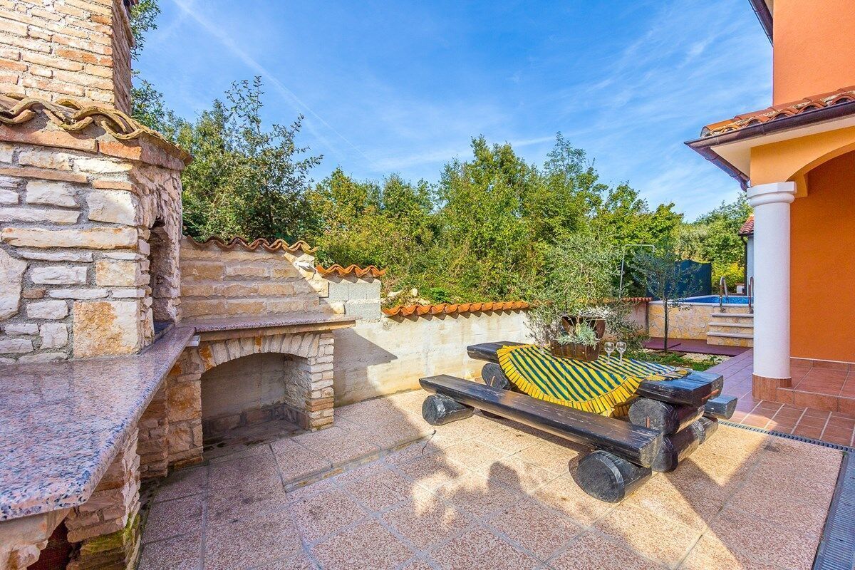 A patio area with a stone barbecue grill, a wooden bench with a striped cushion, and a view of a pool and greenery.