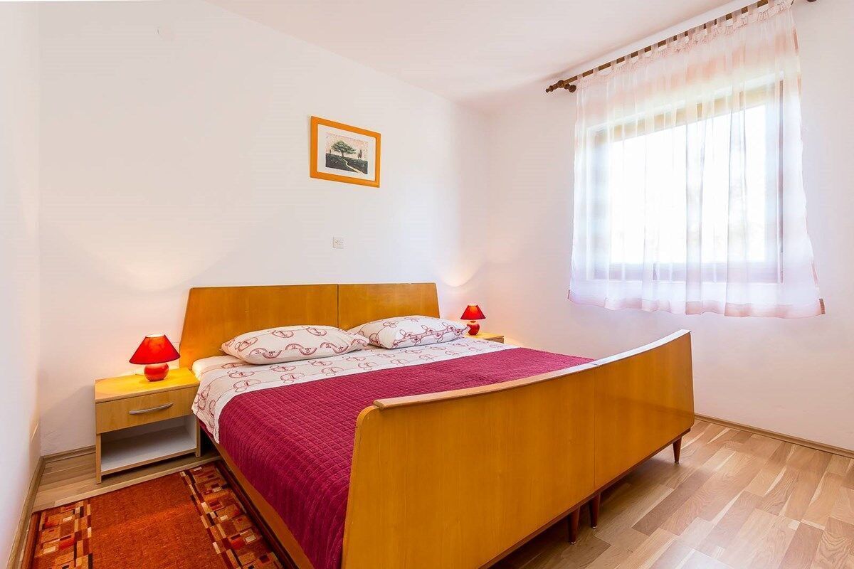 Une chambre à coucher avec un lit en bois, une table de chevet avec une lampe, une commode, une couvre-lit à motifs et une fenêtre avec des rideaux légers.
