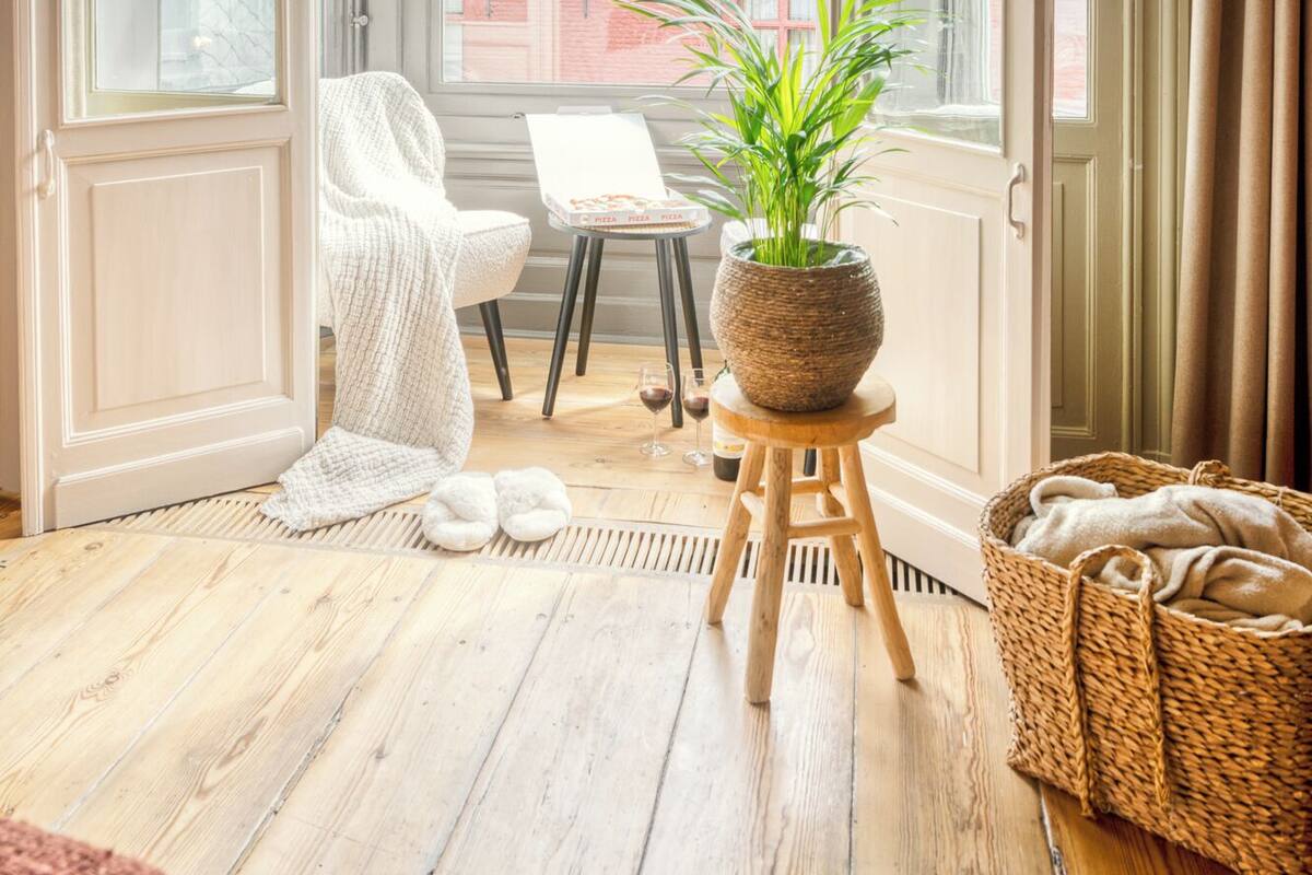 Un balcón abierto con vistas a edificios, una silla blanca, una mesita con planta en maceta y una cesta de mimbre.