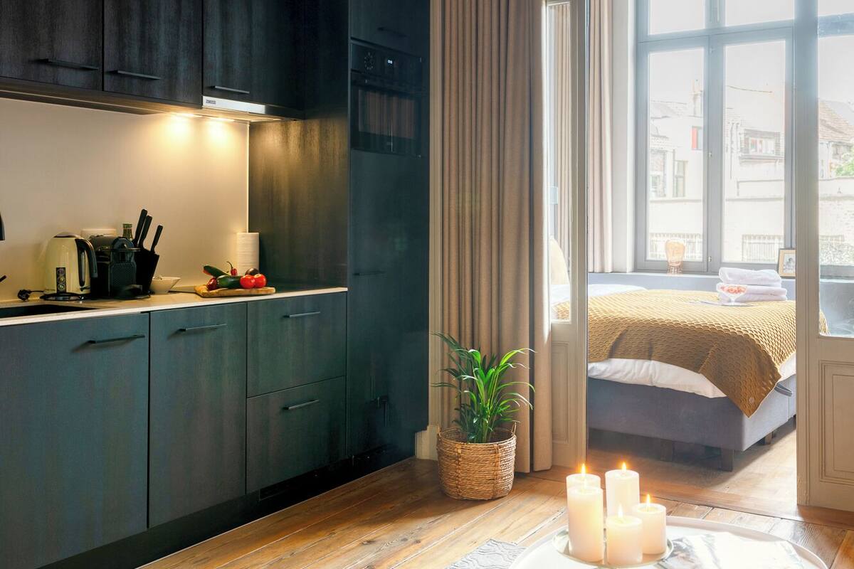 Una cocina moderna con armarios oscuros, horno empotrado y cafetera. Hay un comedor con una mesa redonda y velas. La habitación tiene un ventanal con cortinas y vistas a la ciudad.