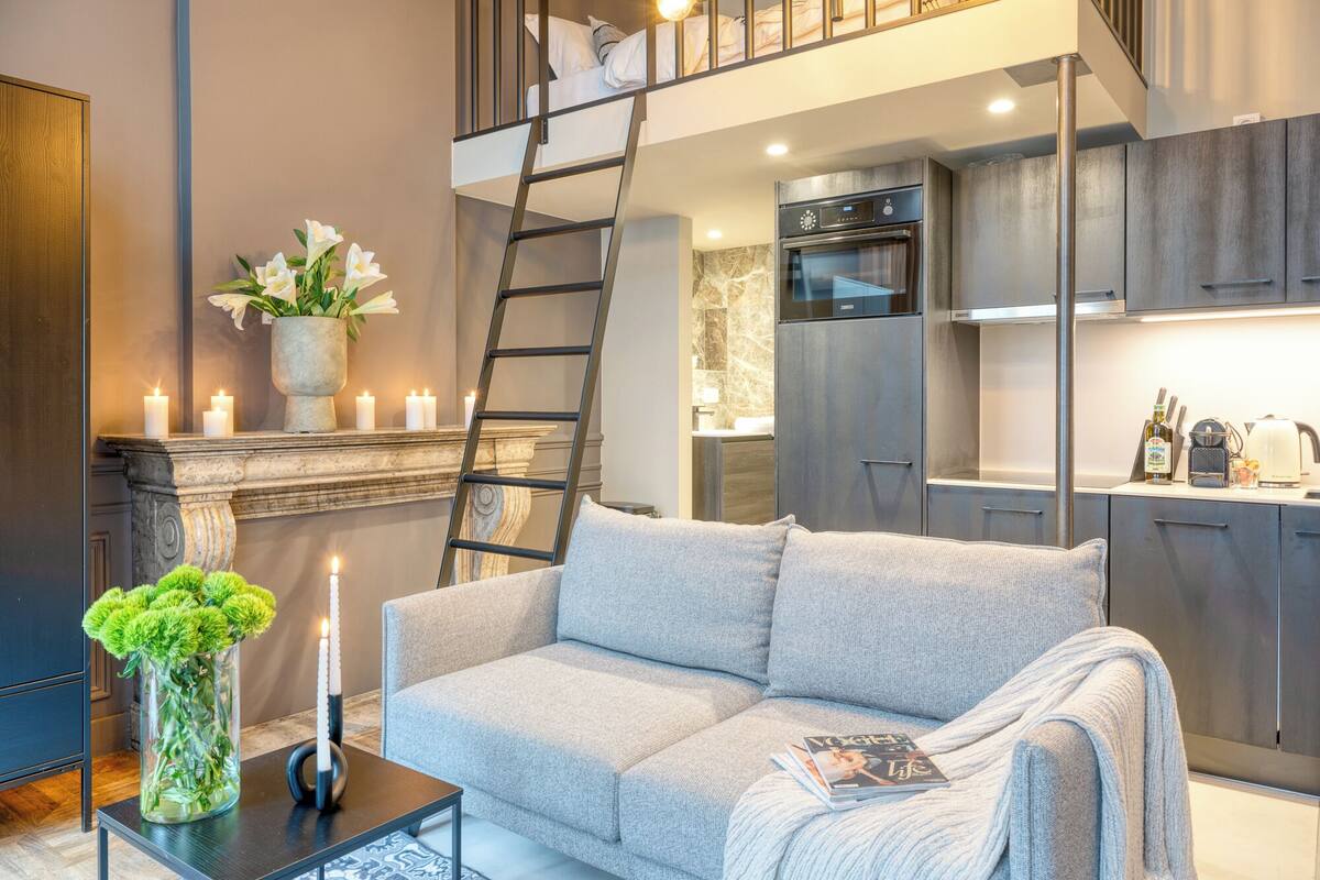 Una sala moderna con un sofá gris, una chimenea con velas, un jarrón con flores y una escalera que lleva a un dormitorio en un altillo.