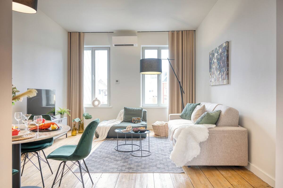 Una sala de estar moderna con un sofá, una mesa de comedor con sillas, un televisor y un gran ventanal con cortinas.