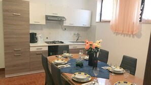 A dining area with a wooden table set for four, white cabinets, a gas stove, and a window with a peach curtain. - Apartments Winny (73623-A1) - Banjole (Banjole)