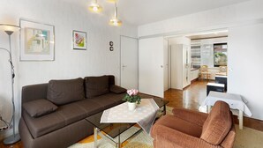 Een moderne woonkamer met een bruine bank, een glazen salontafel en een klein tafeltje met een witte tafelkleed. Boven de tafel hangen twee hanglampen, en aan de muur hangt een ingelijst schilderij.